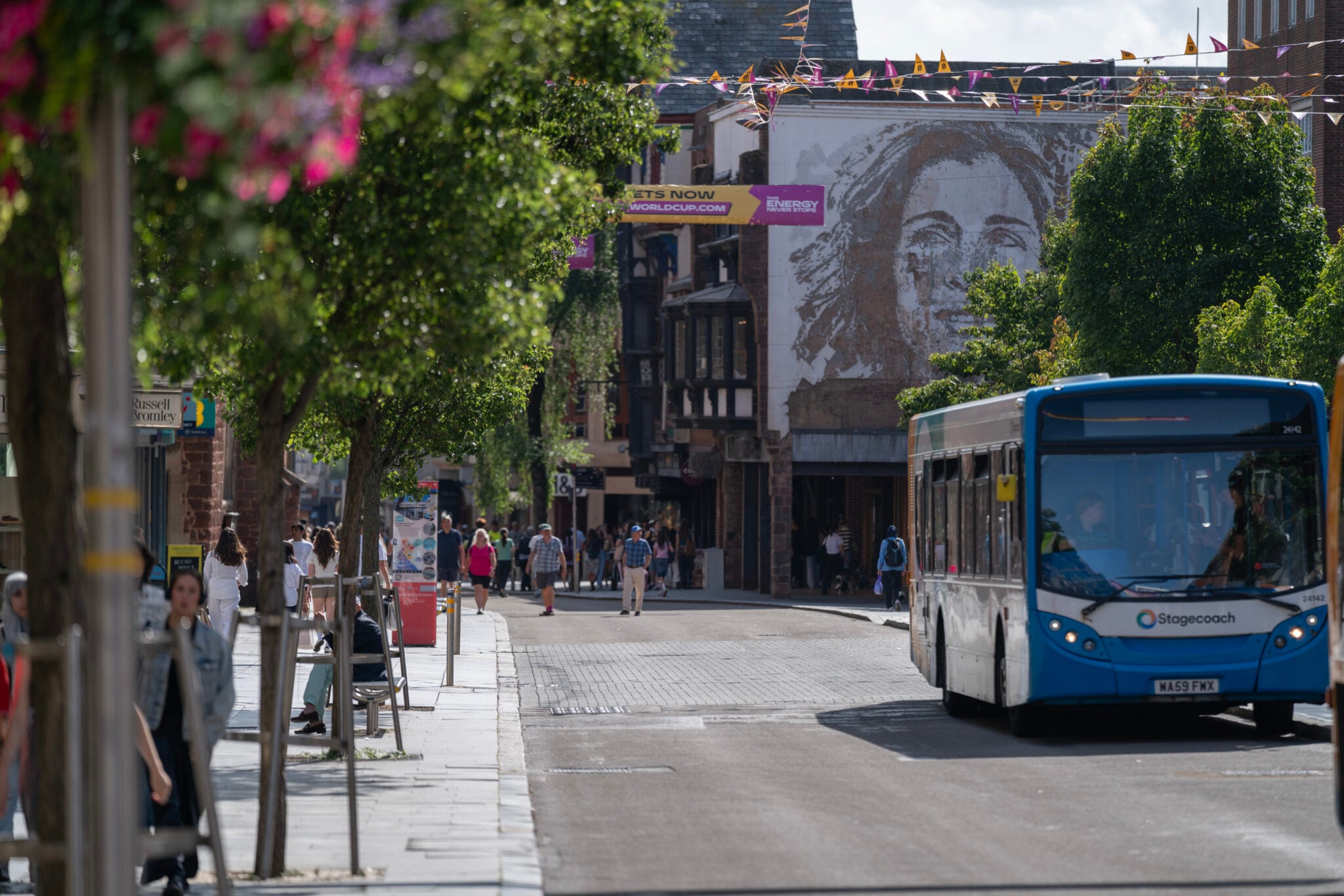 Sunny city street with a blue Stagecoach bus on a cobblestone road, pedestrians walking along tree-lined sidewalks, historic timber-framed buildings in the background, and colourful banners overhead.