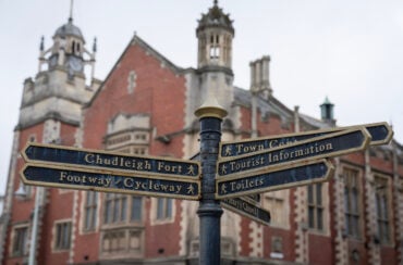 A black metal signpost with several narrow directional signs pointing in different directions. The signs read: “Chudleigh Fort,” “Footway / Cycleway,” “Town Centre,” “Tourist Information,” “Toilets,” and “St. Mary’s Church.” In the background is a large historic red‑brick building with decorative stone detailing, tall windows, and ornate towers.