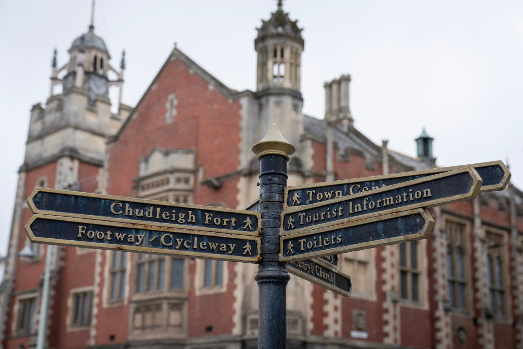 A black metal signpost with several narrow directional signs pointing in different directions. The signs read: “Chudleigh Fort,” “Footway / Cycleway,” “Town Centre,” “Tourist Information,” “Toilets,” and “St. Mary’s Church.” In the background is a large historic red‑brick building with decorative stone detailing, tall windows, and ornate towers.