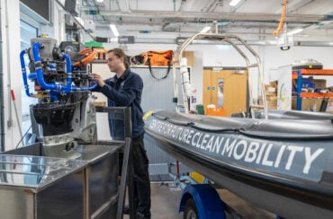A technician works on a marine engine beside a research boat in a workshop.