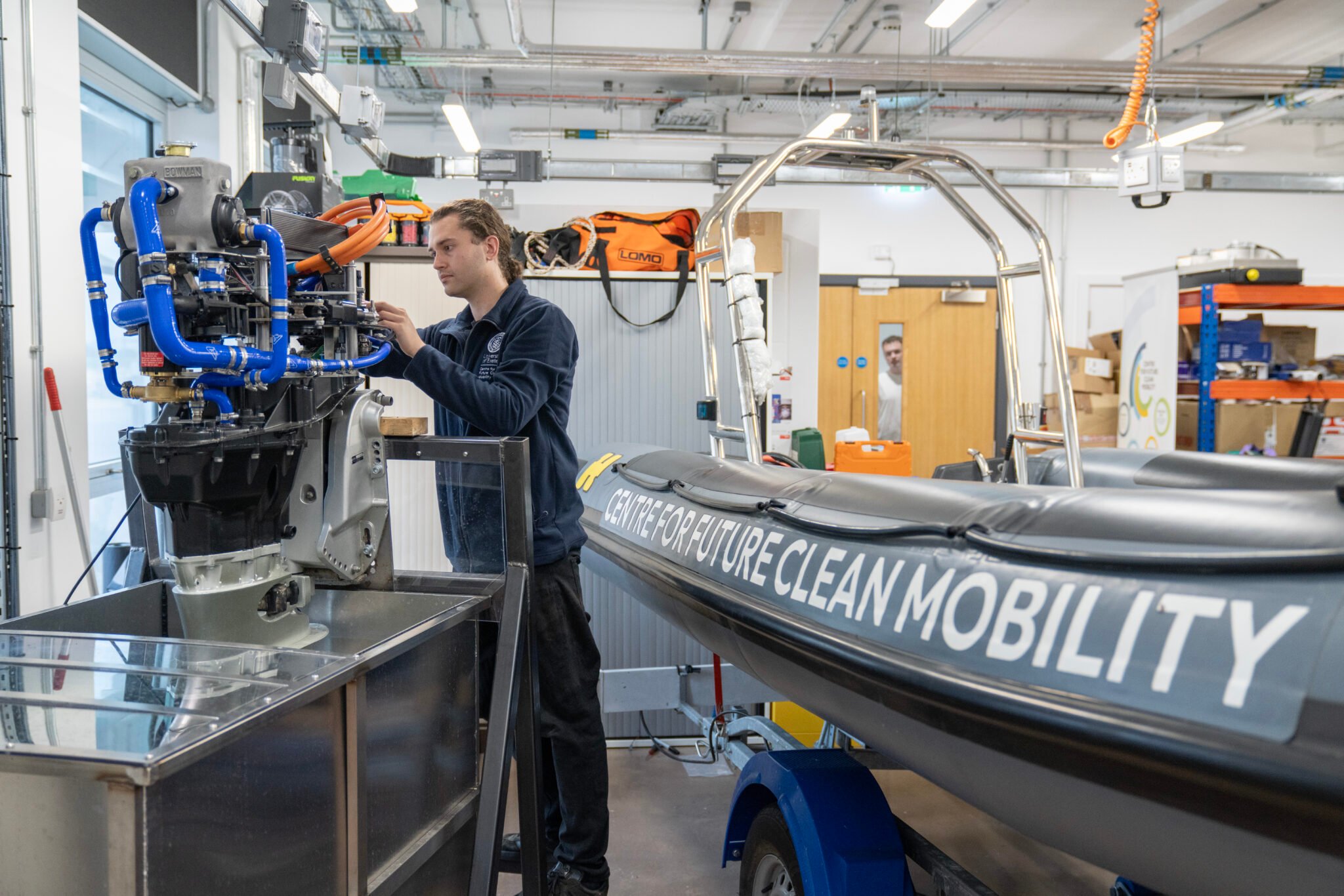 A technician works on a marine engine beside a research boat in a workshop.