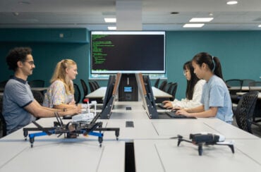 A classroom workspace where four people sit at computers facing each other, with drones on the tables and a large screen displaying lines of code in the background.