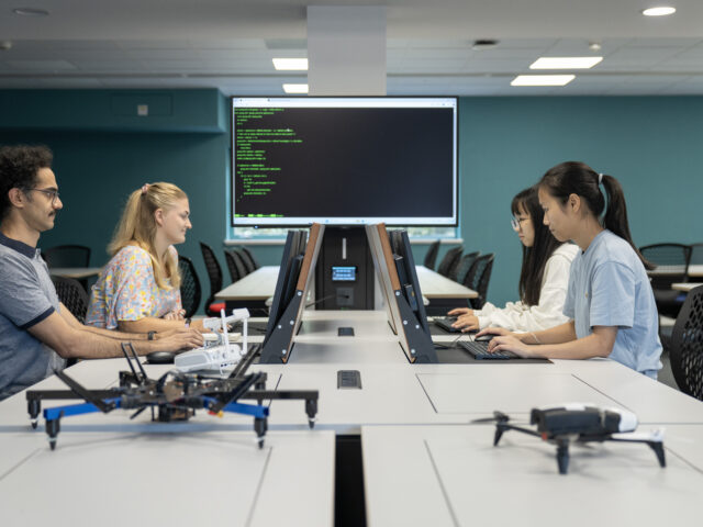 A classroom workspace where four people sit at computers facing each other, with drones on the tables and a large screen displaying lines of code in the background.