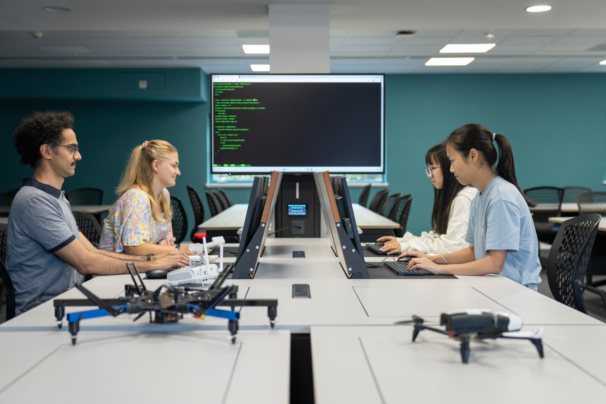 A classroom workspace where four people sit at computers facing each other, with drones on the tables and a large screen displaying lines of code in the background.