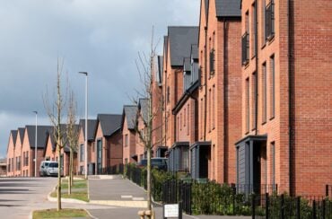 Row of modern red-brick houses along a residential street with young trees, parked cars, and a cloudy sky