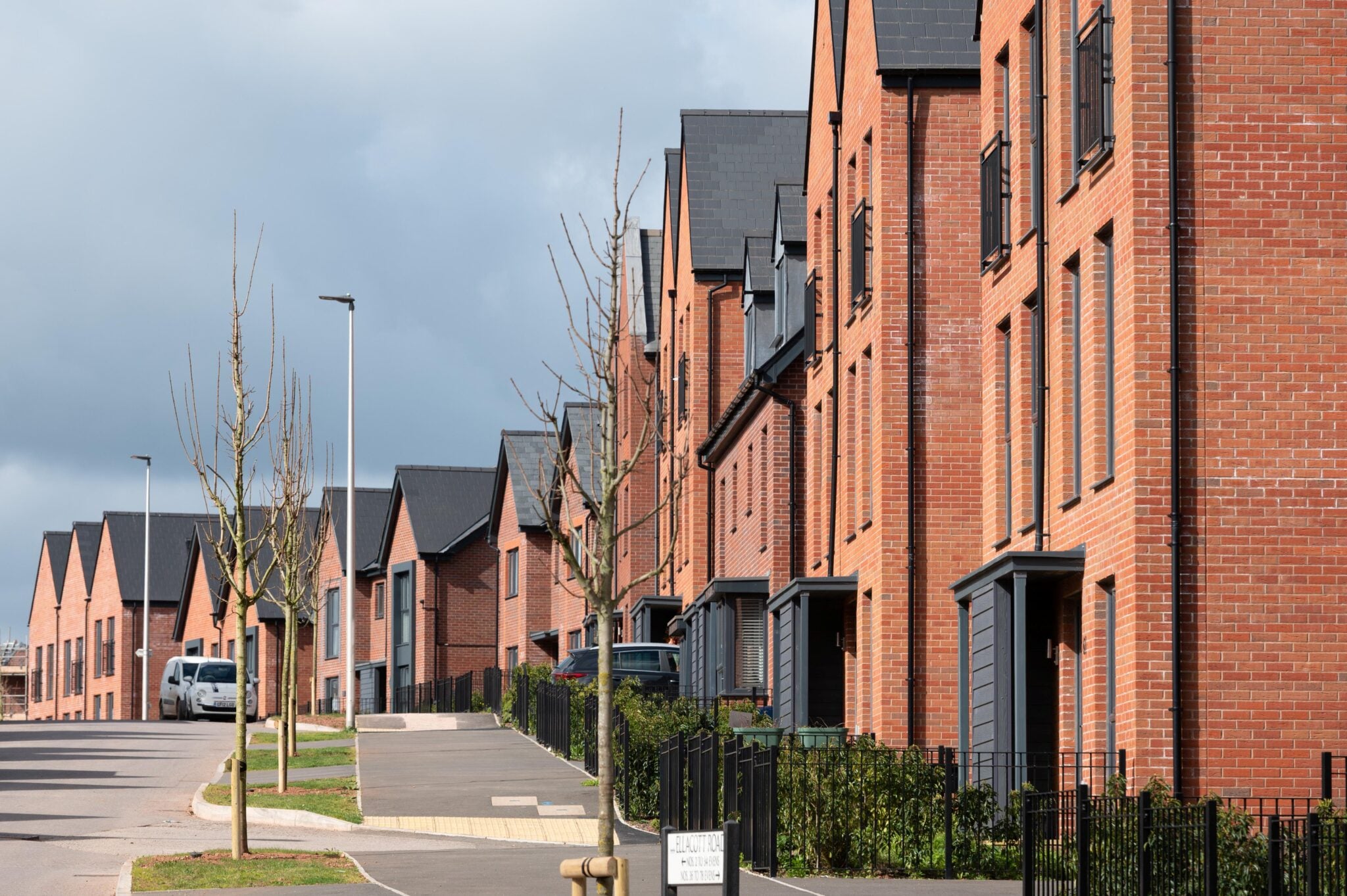 Row of modern red-brick houses along a residential street with young trees, parked cars, and a cloudy sky