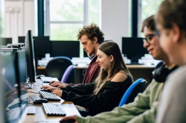three students sat in a computer suite typing on keyboards.