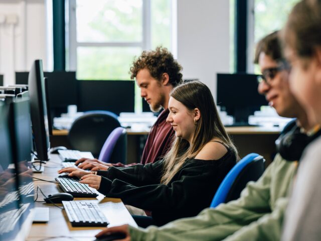three students sat in a computer suite typing on keyboards.