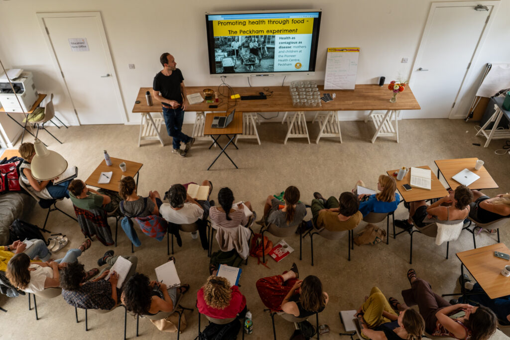 Overhead view of an educational workshop, with participants seated in a circle listening to a presenter standing beside a screen displaying a slide about food and health. Attendees take notes while tables, jars, and workshop materials are arranged around the room.