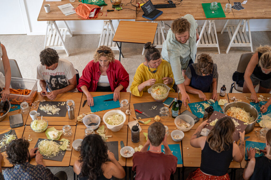 Overhead view of a group cooking workshop, with participants seated and standing around large wooden tables preparing vegetables. Chopping boards, knives, bowls of shredded cabbage and sliced onions, jars, and kitchen equipment are spread across the tables in a bright indoor space.