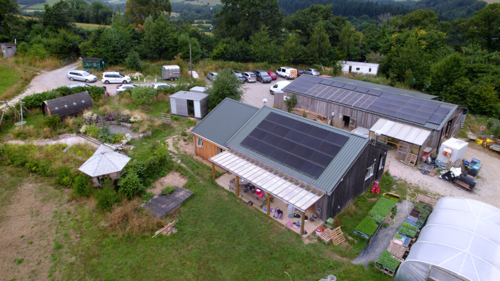 Aerial view of the Apricot Centre featuring wooden buildings with large rooftop solar panels, surrounded by gardens, growing areas, and trees. Several vehicles are parked nearby, with rolling countryside in the background.