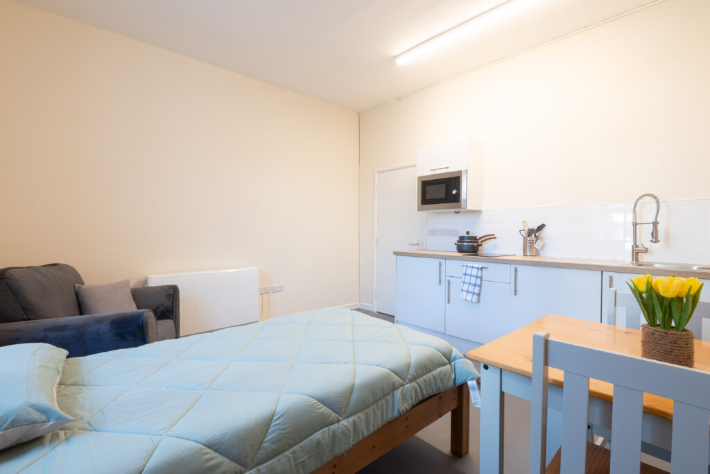 Studio-style room with a bed, armchair and compact kitchenette, furnished in a simple, modern style.