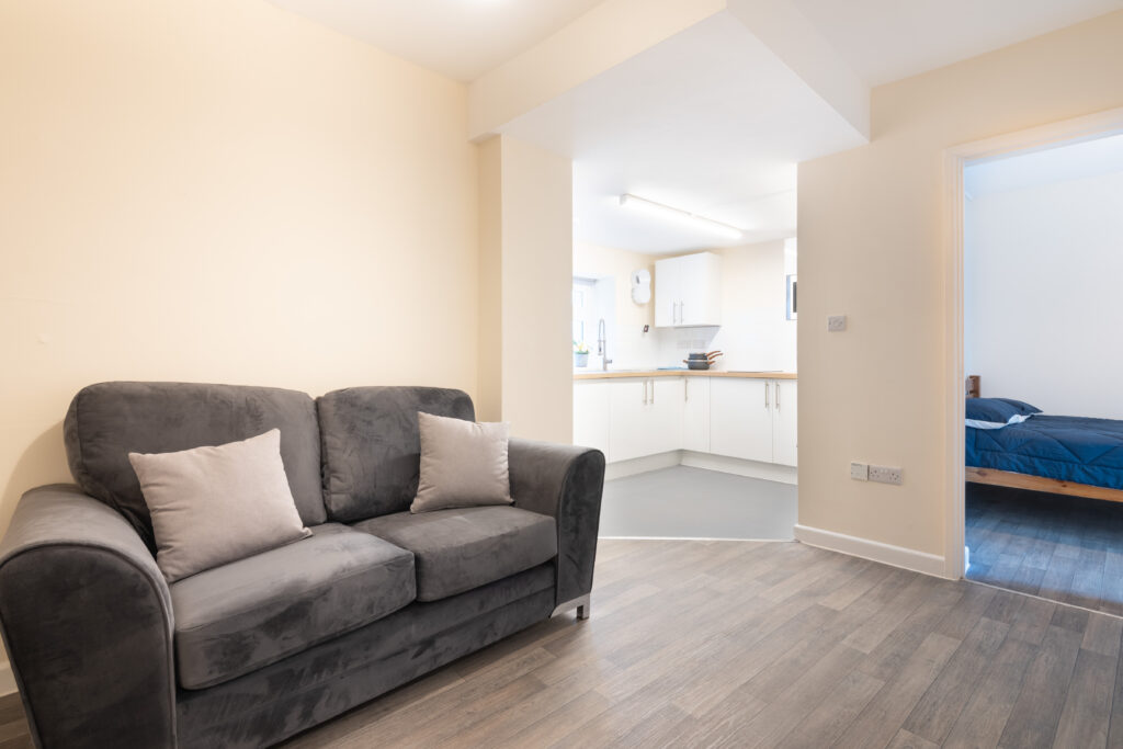 Open-plan apartment with a grey sofa, fitted kitchen and separate bedroom visible through an open doorway.