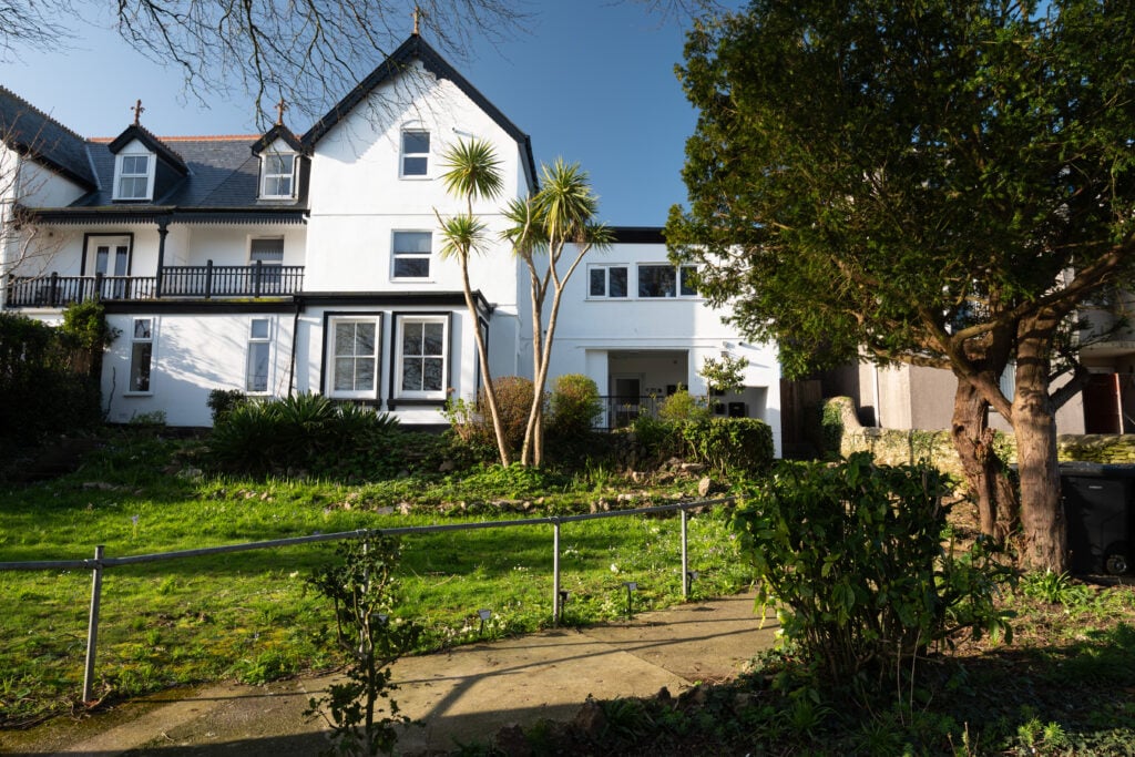 White multi-storey residential building with balconies and large windows, set behind a landscaped garden with mature trees and lawn on a sunny day.