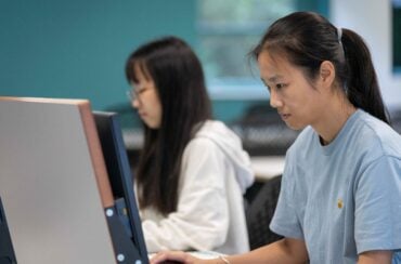 Two people seated at desktop computers in a computer lab or office setting, focused on work. The person in the foreground wears a light blue T-shirt, while another person is visible in the background, with the image softly blurred behind them.