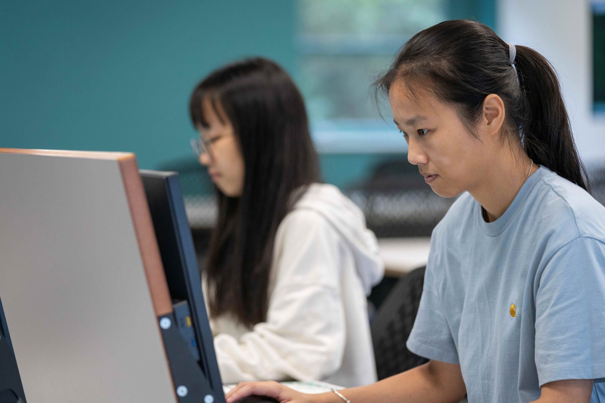 Two people seated at desktop computers in a computer lab or office setting, focused on work. The person in the foreground wears a light blue T-shirt, while another person is visible in the background, with the image softly blurred behind them.
