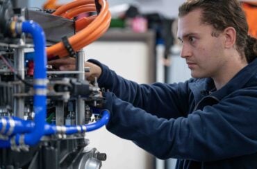 A technician works on a piece of industrial machinery, adjusting components among blue and orange pipes and cables in a workshop setting.