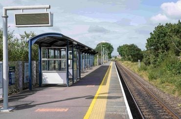 A railway station platform with a covered waiting area, digital information display and a single railway track, surrounded by trees and greenery.
