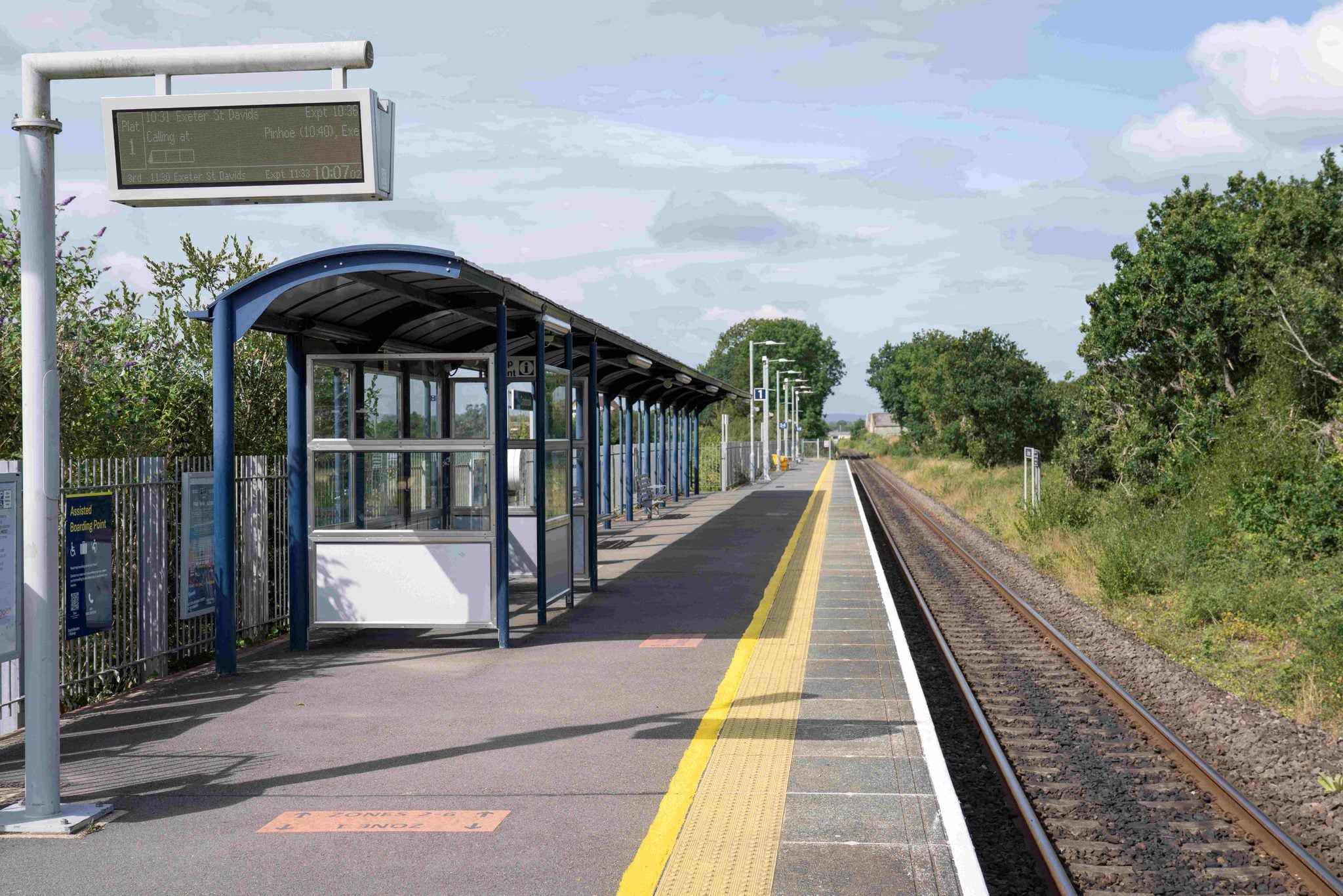 A railway station platform with a covered waiting area, digital information display and a single railway track, surrounded by trees and greenery.
